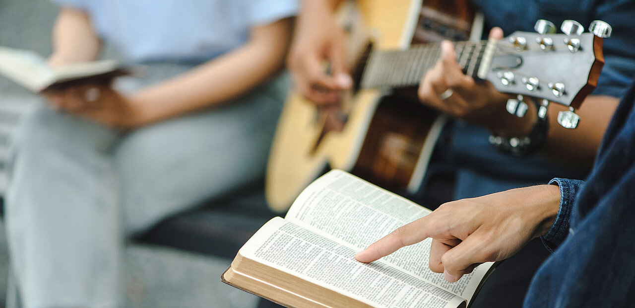 Jemand spielt Gitarre und zwei Personen lesen die Bibel