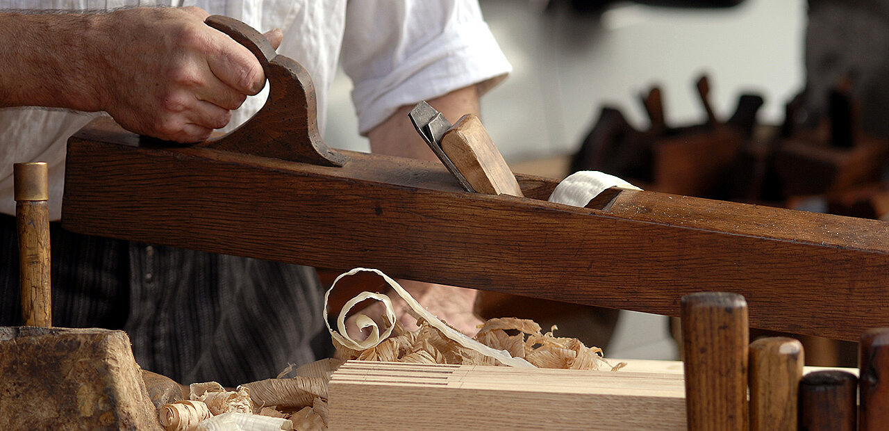 Ein Tischler bearbeitet mit einem Handhobel ein Stück Holz.