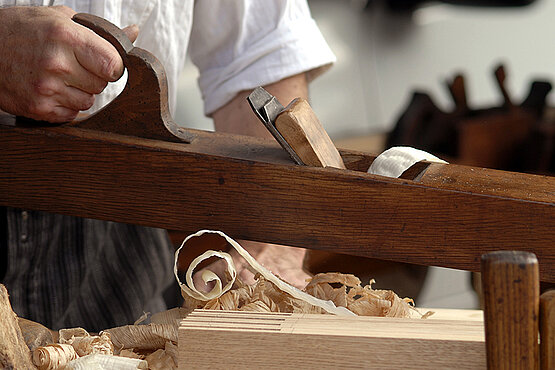 Ein Tischler bearbeitet mit einem Handhobel ein Stück Holz.