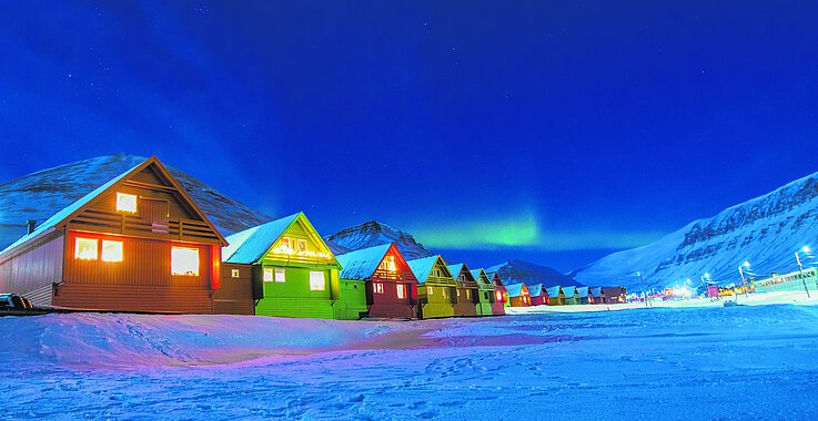 Fast ein wenig unwirklich wirkt die Stadt Longyearbyen, der norwegische Hauptort der Inselgruppe von Spitzbergen, wenn er in der Polarnacht Tag und Nacht nur von den künstlichem Licht erleuchtet wird. Am Horizont sieht man Polarlichter tanzen.