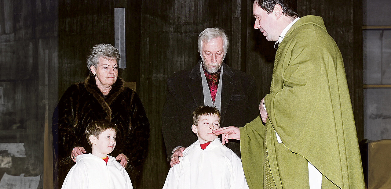 Zwei Kinder werden getauft. Am Bild zu sehen die Kinder,  und deren Großeltern und der Priester Der Priester berührt den Mund eines der Täuflinge.