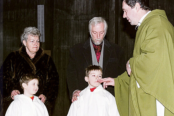 Zwei Kinder werden getauft. Am Bild zu sehen die Kinder,  und deren Großeltern und der Priester Der Priester berührt den Mund eines der Täuflinge.